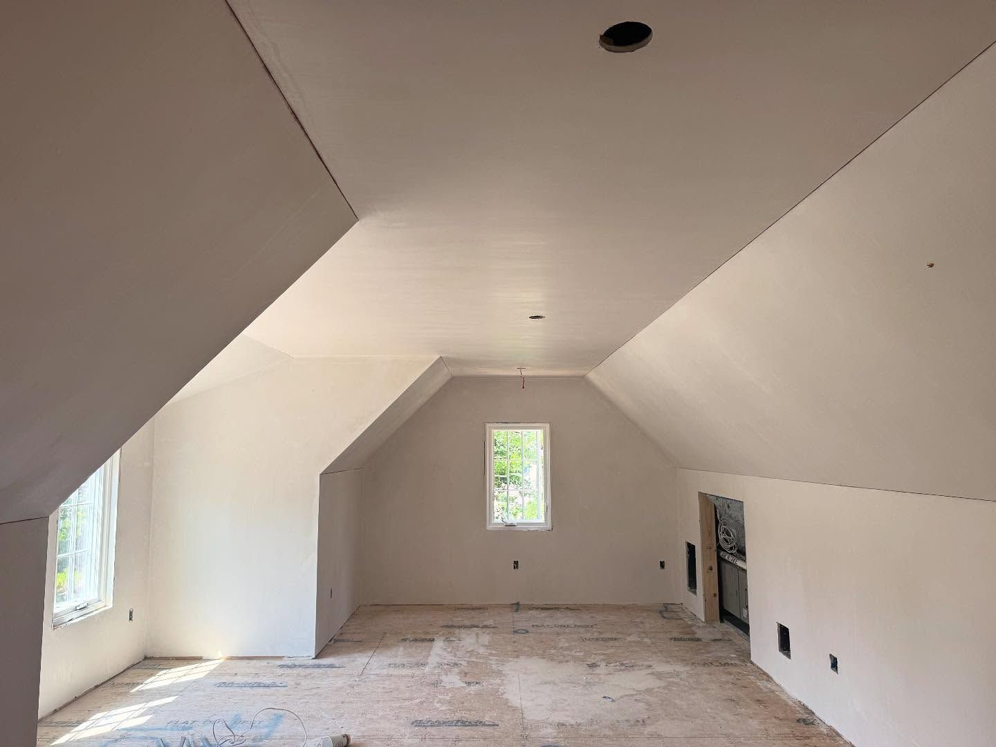 Interior of a room under construction with slanted ceilings, unfinished walls, window, and exposed floor.
