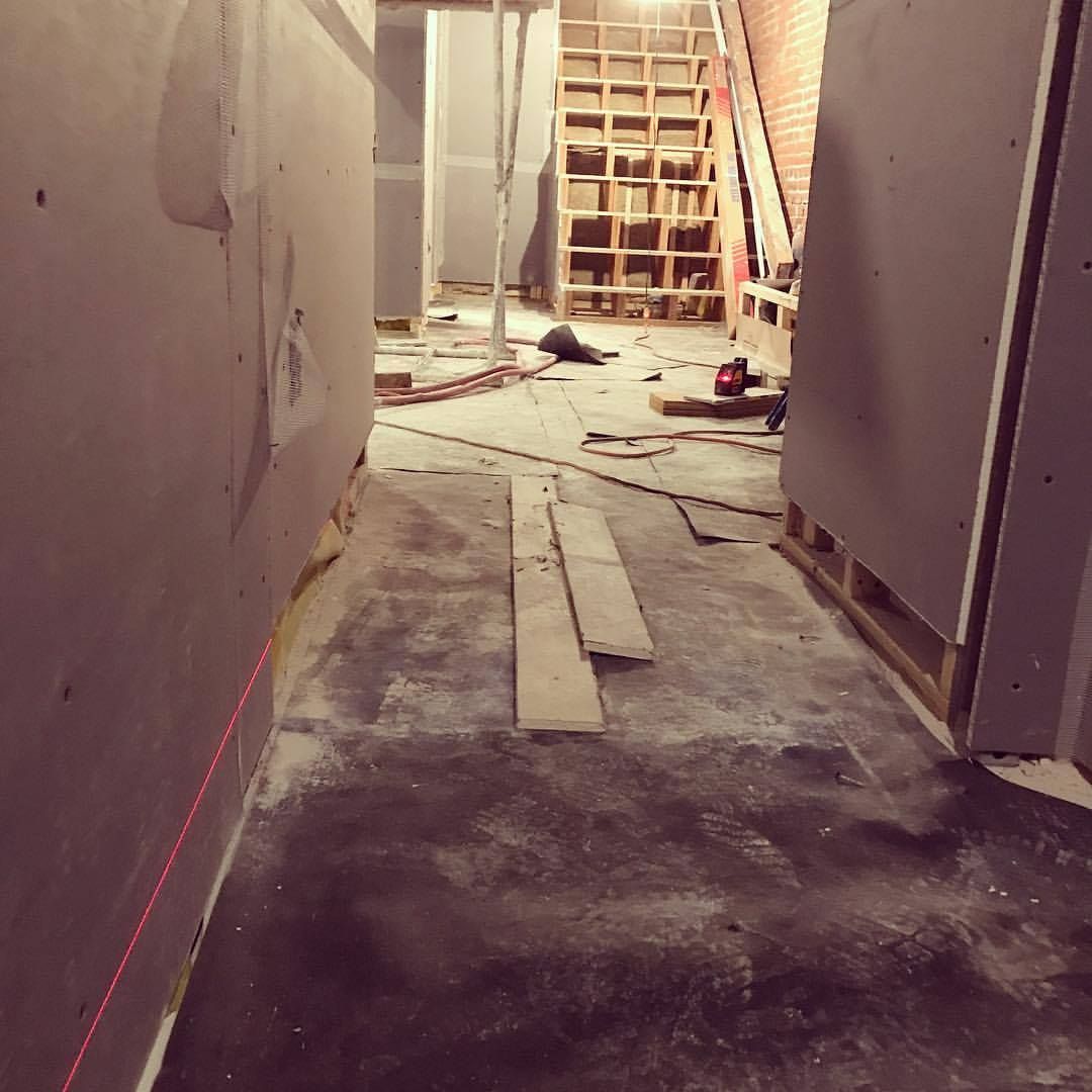 Narrow hallway under construction; gray walls, concrete floor, wood, tools, and ladder visible.