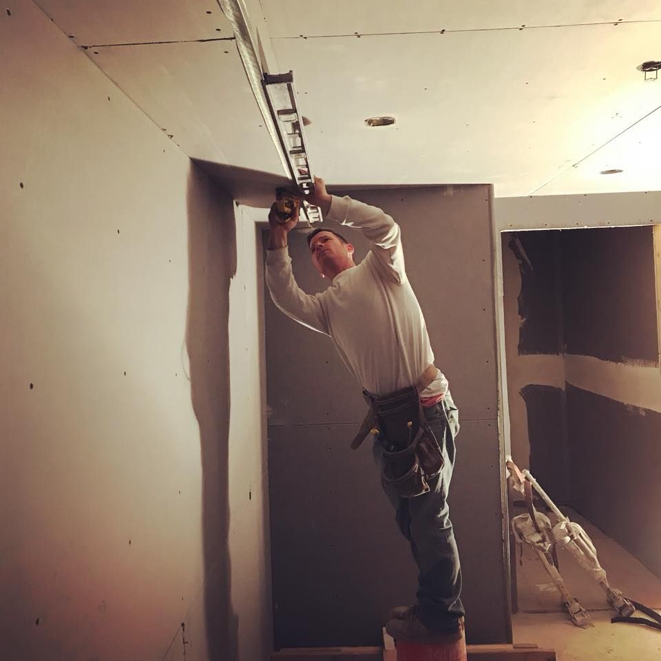 Construction worker on a stool installing a track light in a room with gray walls.