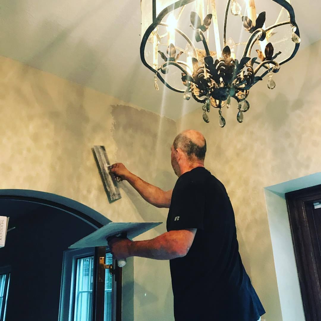 Man plastering a wall indoors, under a chandelier.