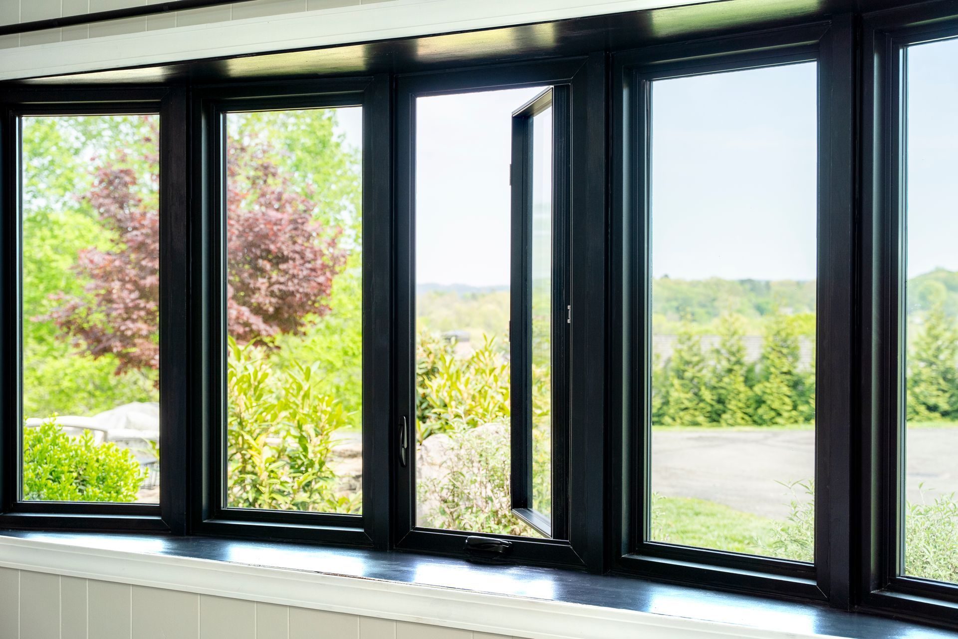 Black-framed windows, one open, overlook a bright green landscape with trees and a blue sky.