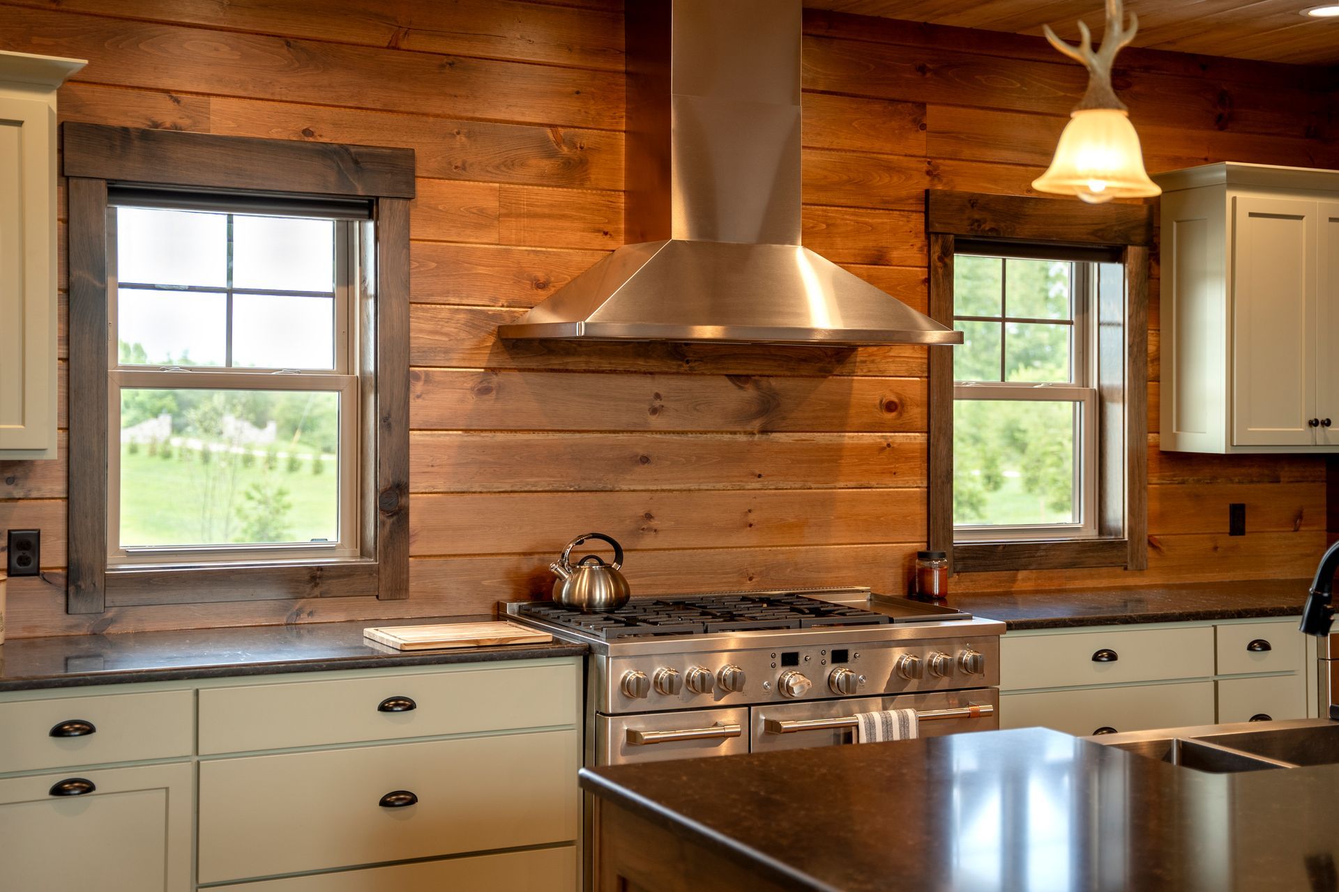 Rustic kitchen with wood paneling, stainless steel range hood, windows, and cream-colored cabinets.