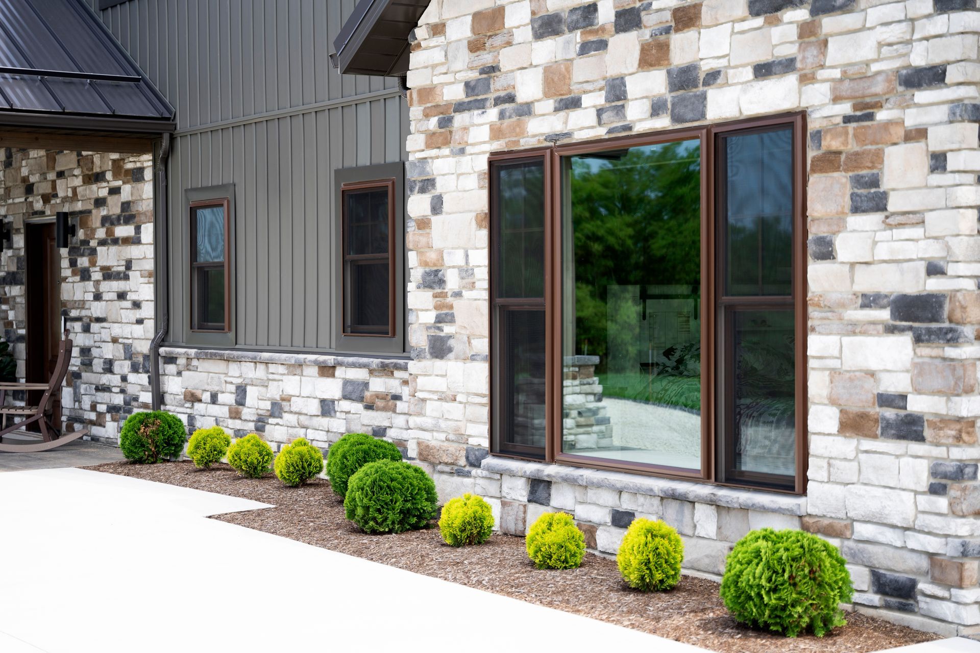 Exterior view of a house with stone and wood siding, windows, and green shrubbery along the sidewalk.
