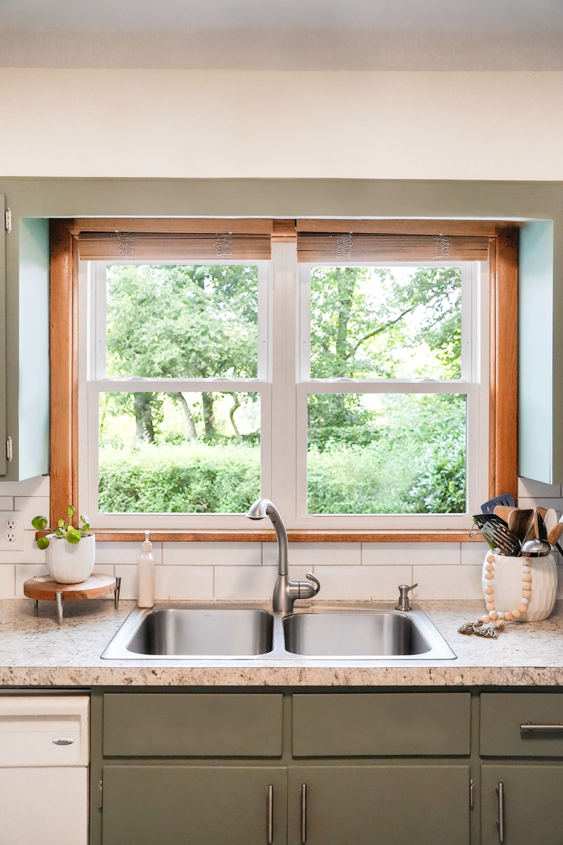 Kitchen sink under a window with a view of greenery; green cabinets, light countertop, wooden accents.
