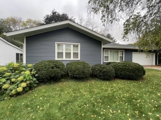 A blue, single-story house with a gabled roof, white trim, a large window, and rounded bushes in the front yard.