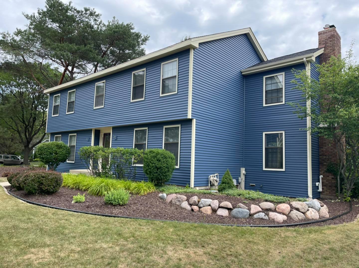 Two-story, blue-sided colonial house with a brick chimney and landscaped front yard featuring rock-lined garden beds.