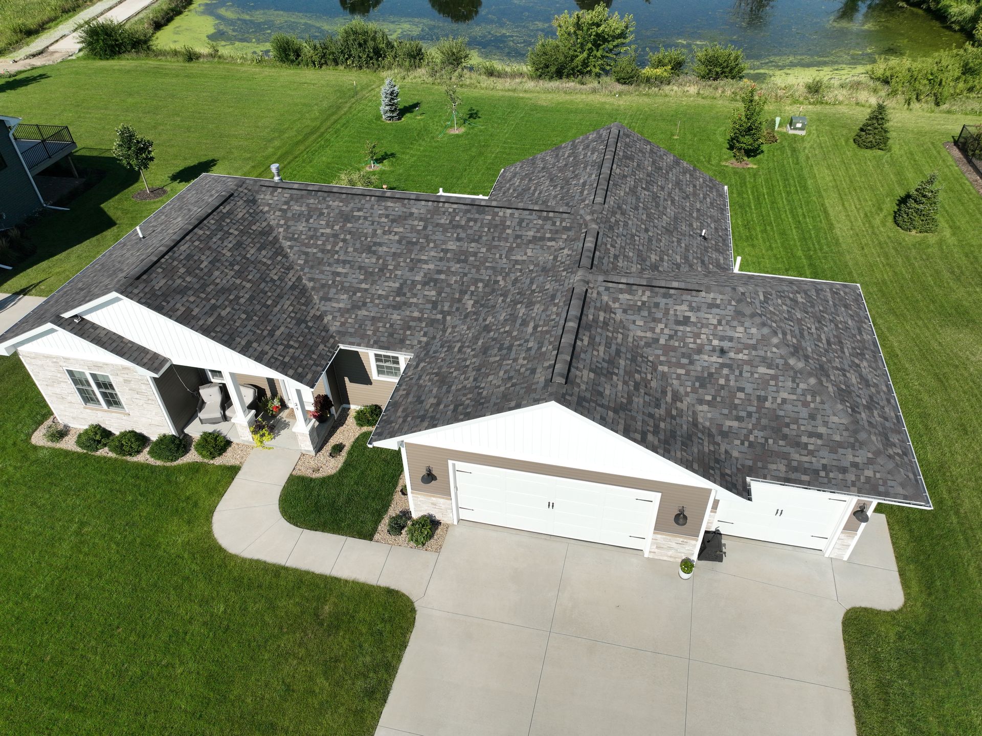 Aerial view of a house with a gray shingled roof, white trim, and a concrete driveway surrounded by a large green lawn.