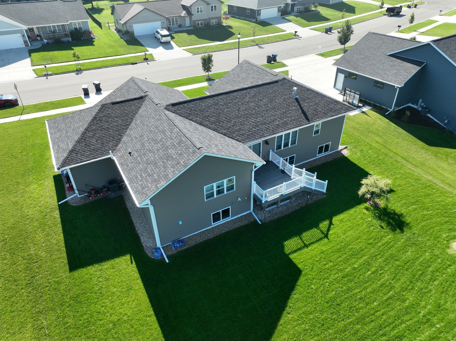 An aerial view of a gray residential house with a dark shingled roof, a back deck, and a green lawn in a suburban area.