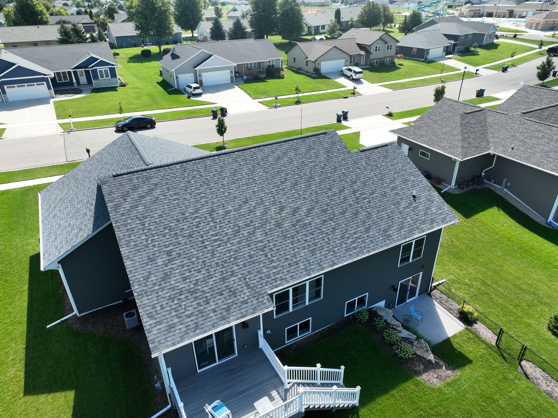 An aerial view of a dark gray house with a shingled roof, a wooden deck, and a green lawn in a suburban neighborhood.