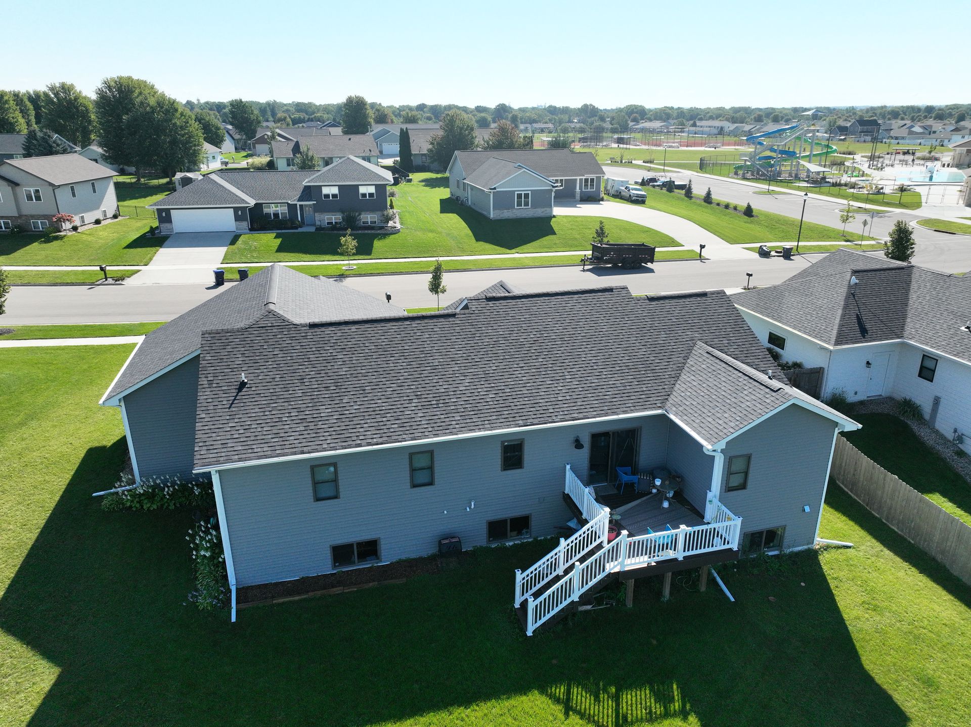 Aerial view of a gray suburban house with a dark shingled roof, a wooden deck, and a green lawn on a sunny day.
