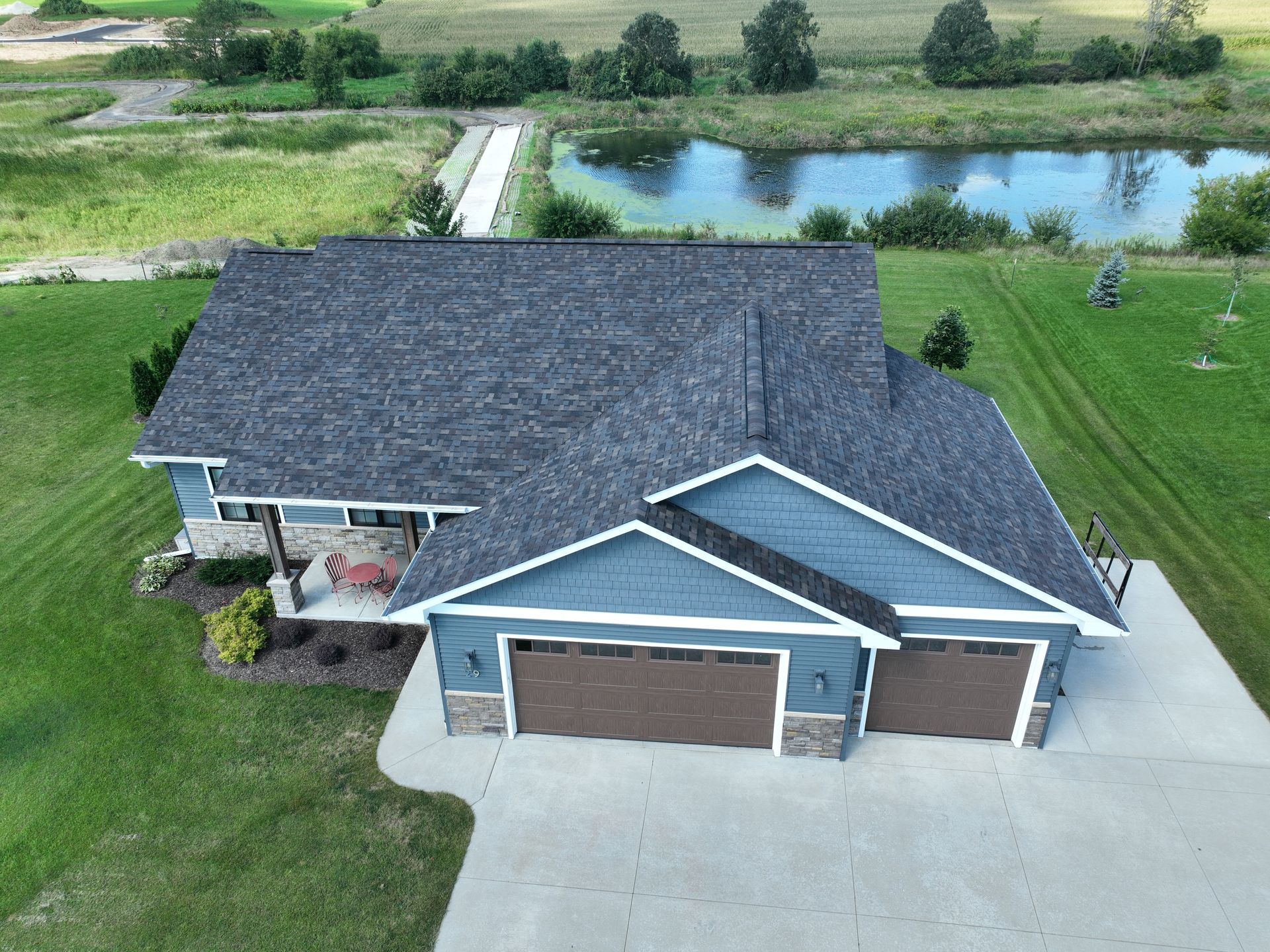 Aerial view of a blue house with brown garage doors and a grey roof, situated on a grassy lot next to a pond.