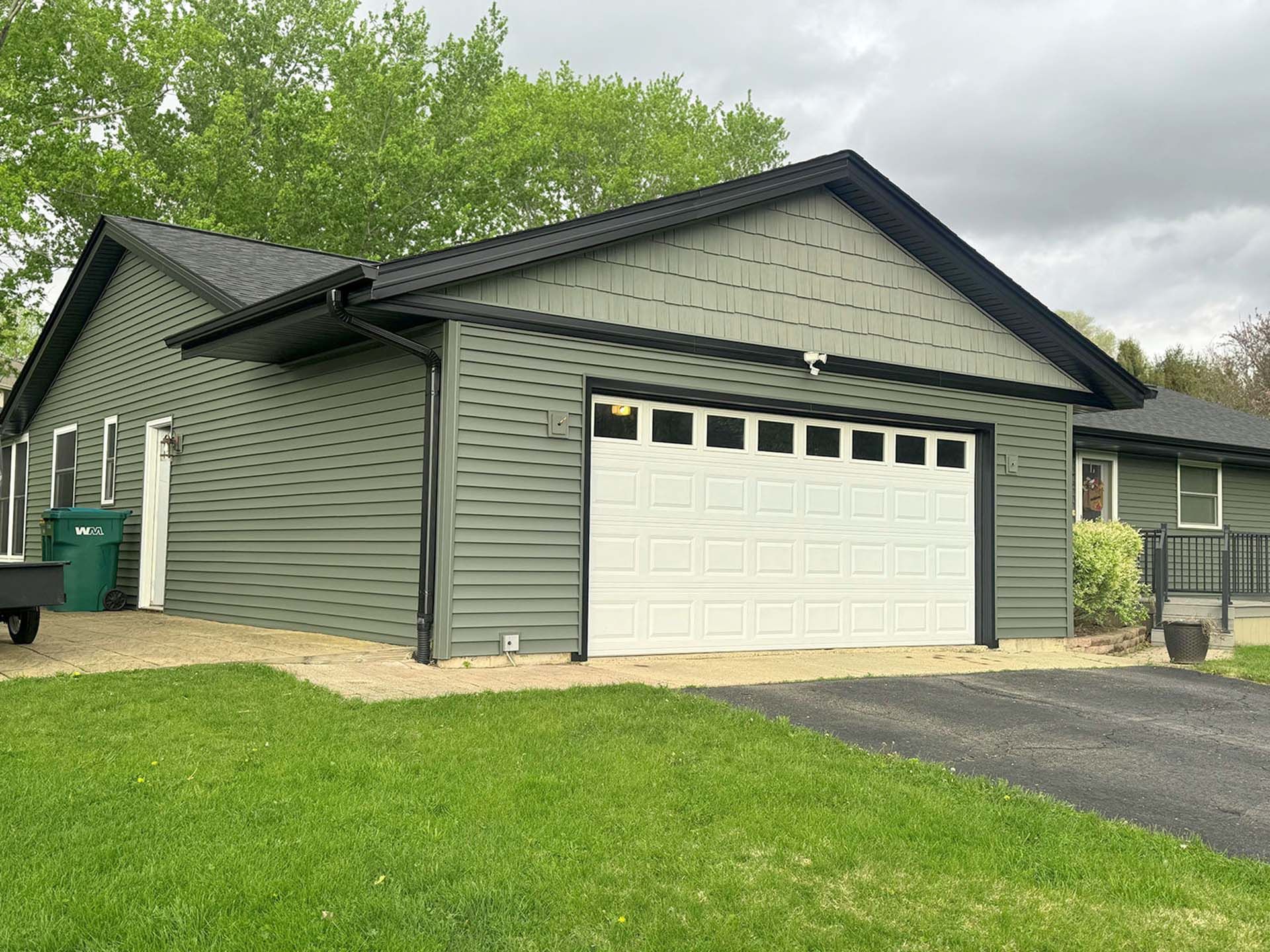 Green house with white garage door, driveway, and lawn under cloudy skies