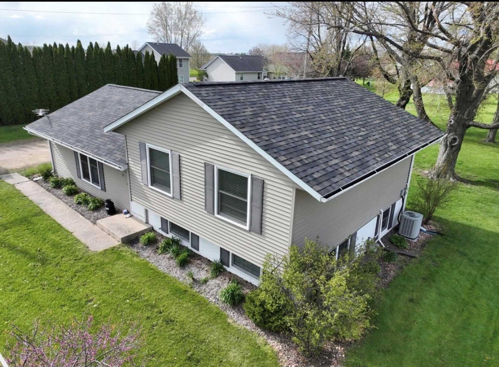 Suburban house with gray siding and dark shingled roof, viewed from above beside a lawn and trees