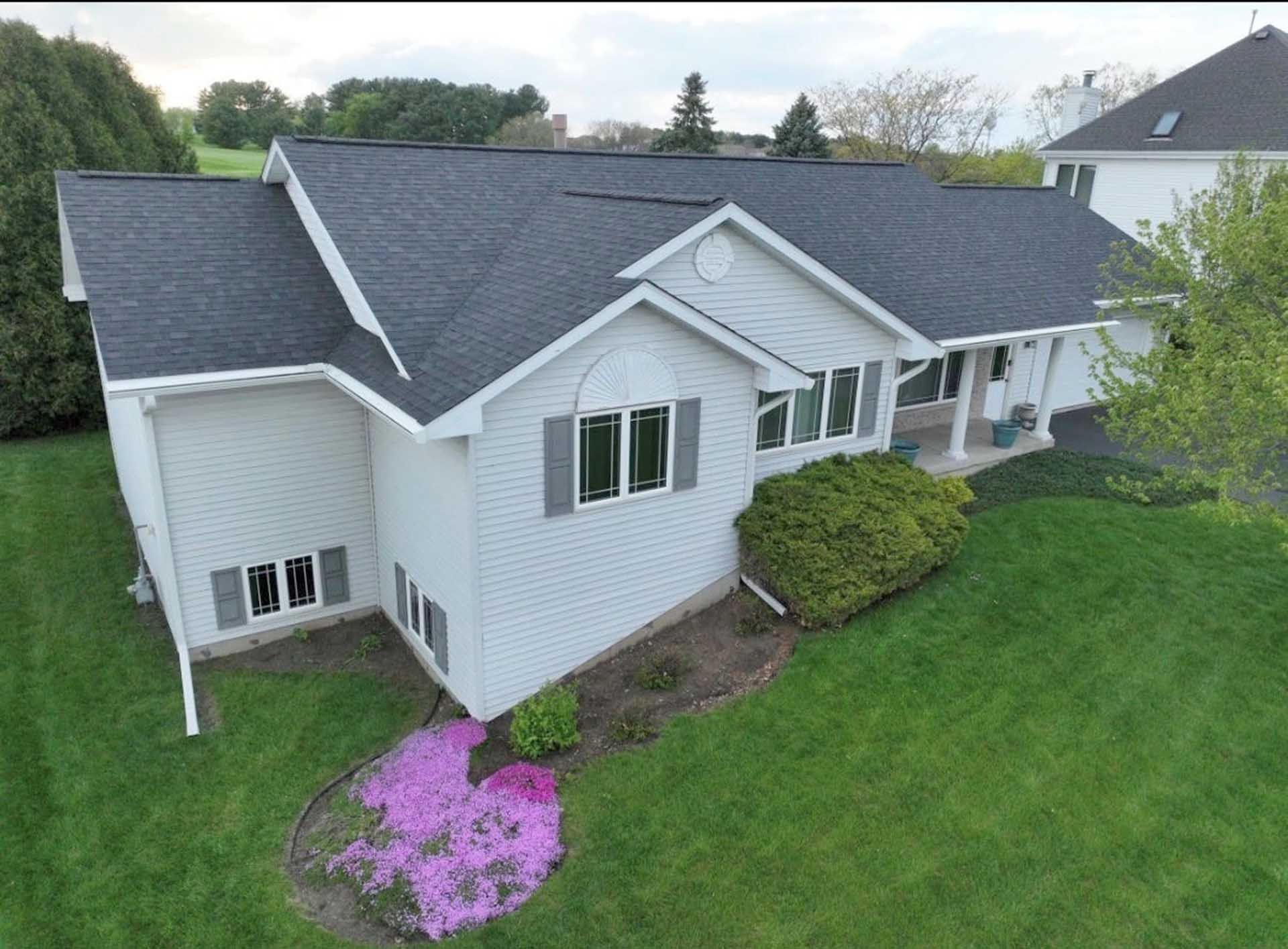 Aerial view of a white suburban house with a dark roof and green lawn, accented by pink flowers