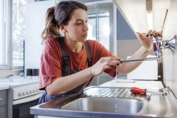 A woman is fixing a faucet in a kitchen with a screwdriver.