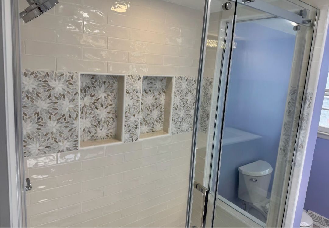 Shower with glass doors and tiled walls. Recessed shelf area with decorative tile. White and grey.