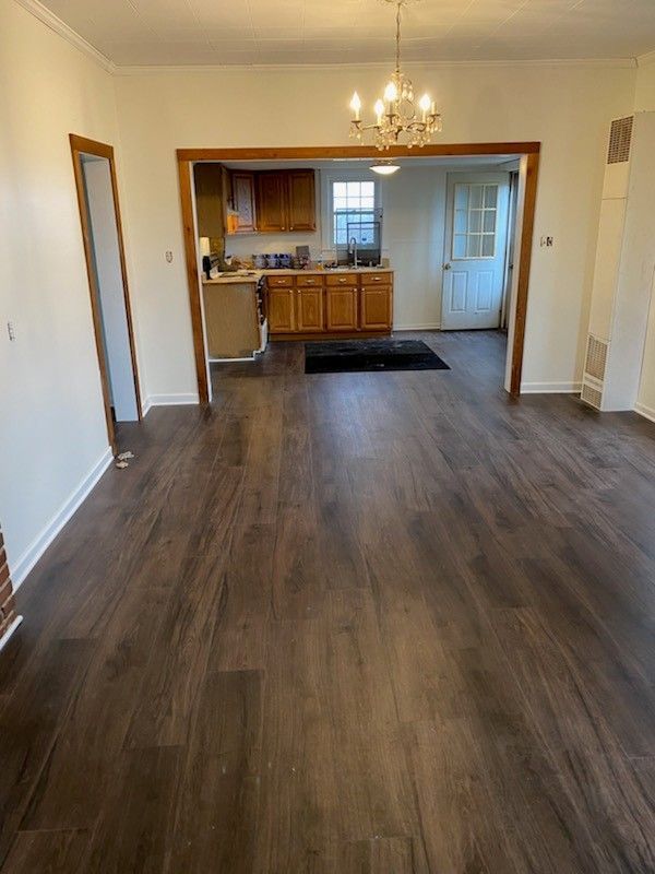 A wide-angle view of a room with dark wood flooring leading through an open doorway into a kitchen with wood cabinets.