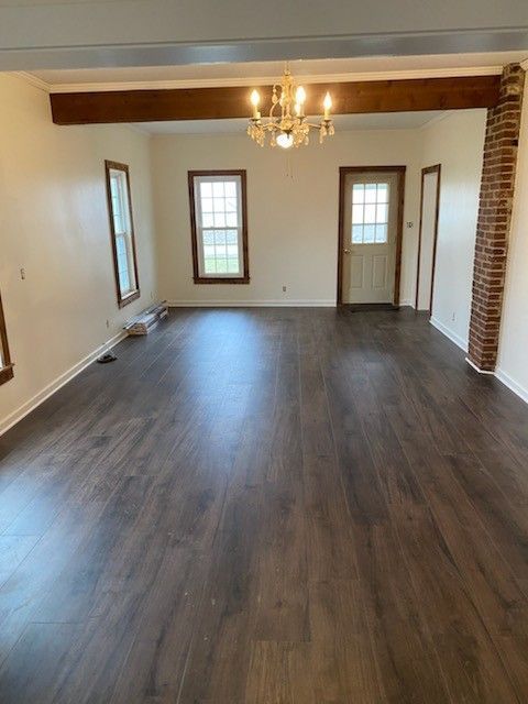 A spacious, empty living room with dark wood-style flooring, light walls, a brick pillar, and a central chandelier.