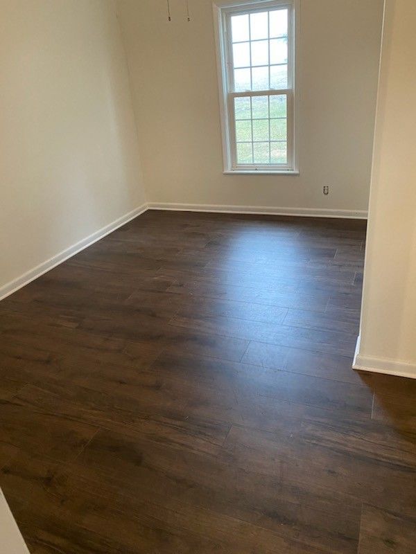 A bright, empty room with dark wood-look laminate flooring and white walls, featuring a single window.