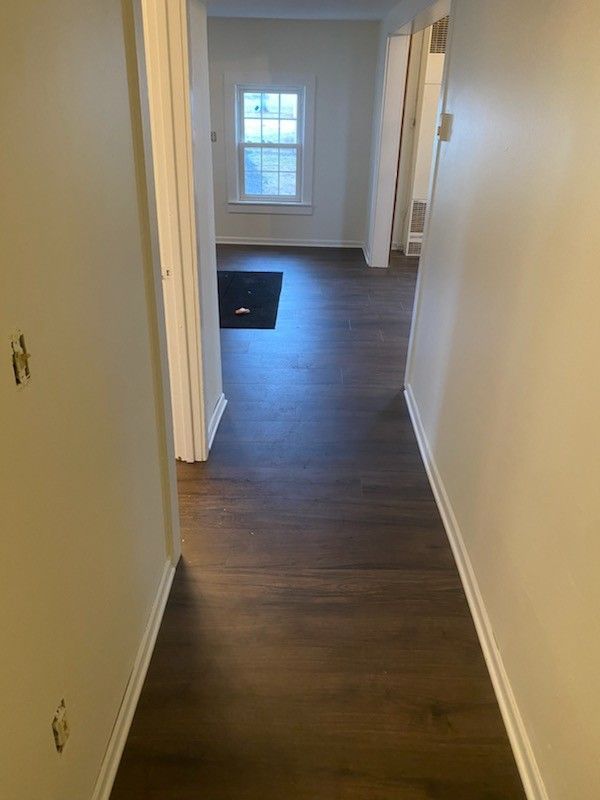 A view down a neutral-walled hallway with dark wood-look flooring leading toward a windowed room with a black floor mat.