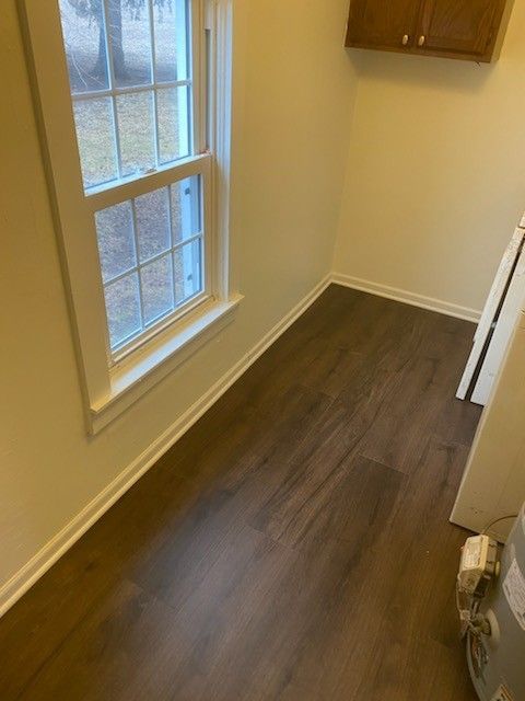 A laundry room corner with dark wood-look floors, cream walls, a multi-pane window, and a wooden upper cabinet.