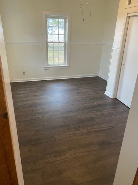 An empty room with wood-look laminate flooring, cream-colored walls with wainscoting, and a window centered on the wall.