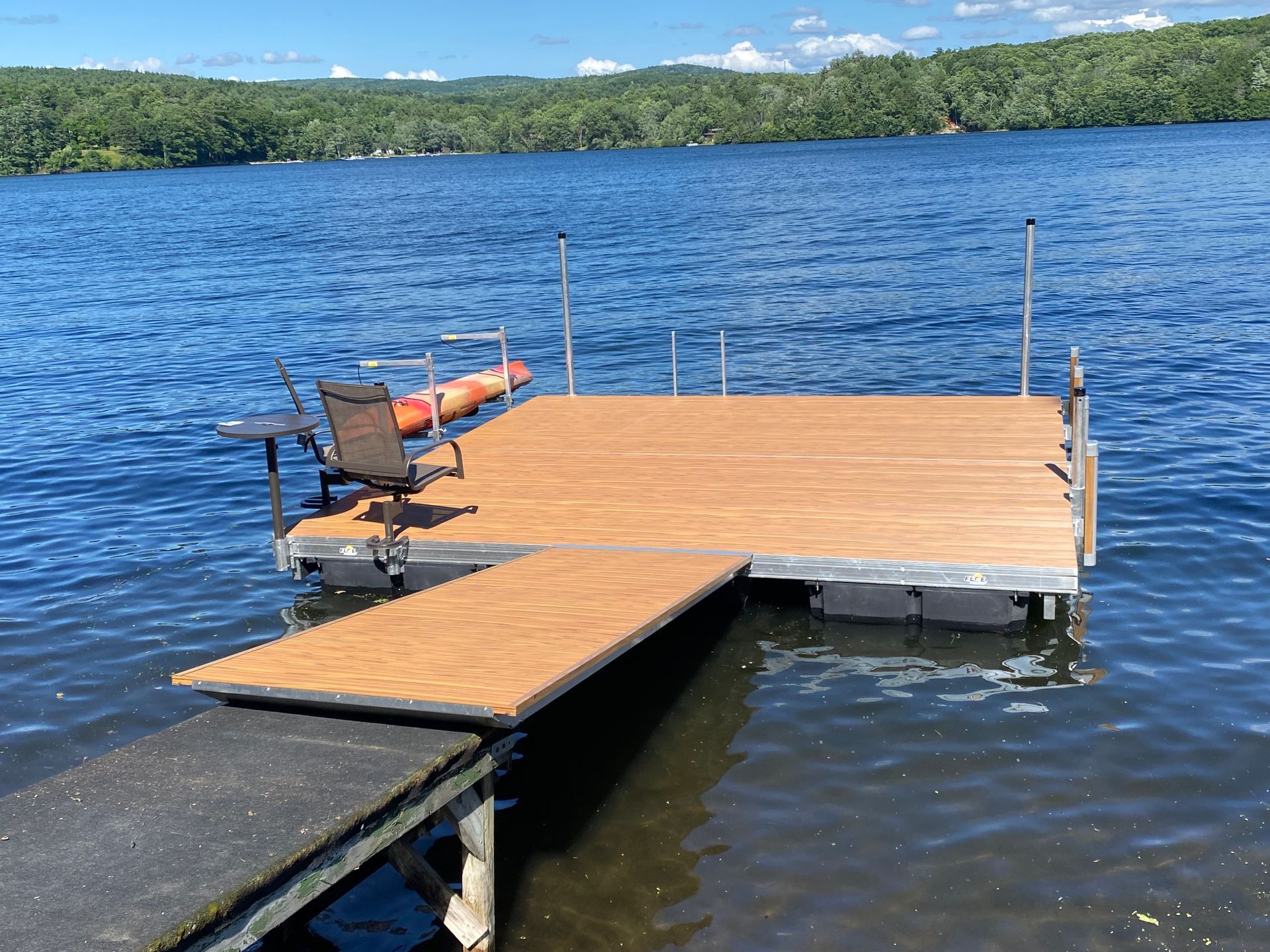 Wooden dock with a chair on a calm blue lake, facing a wooded shoreline under a clear sky