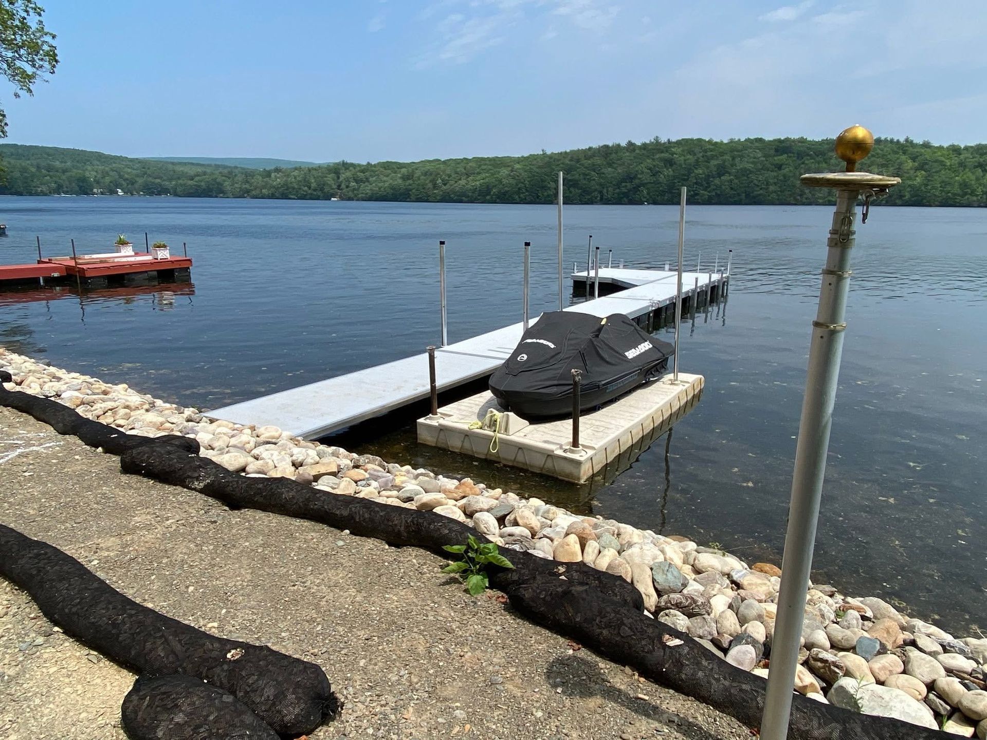 A lakefront scene with a dock and a jet ski with a black cover, viewed from a rocky shoreline under a clear blue sky.