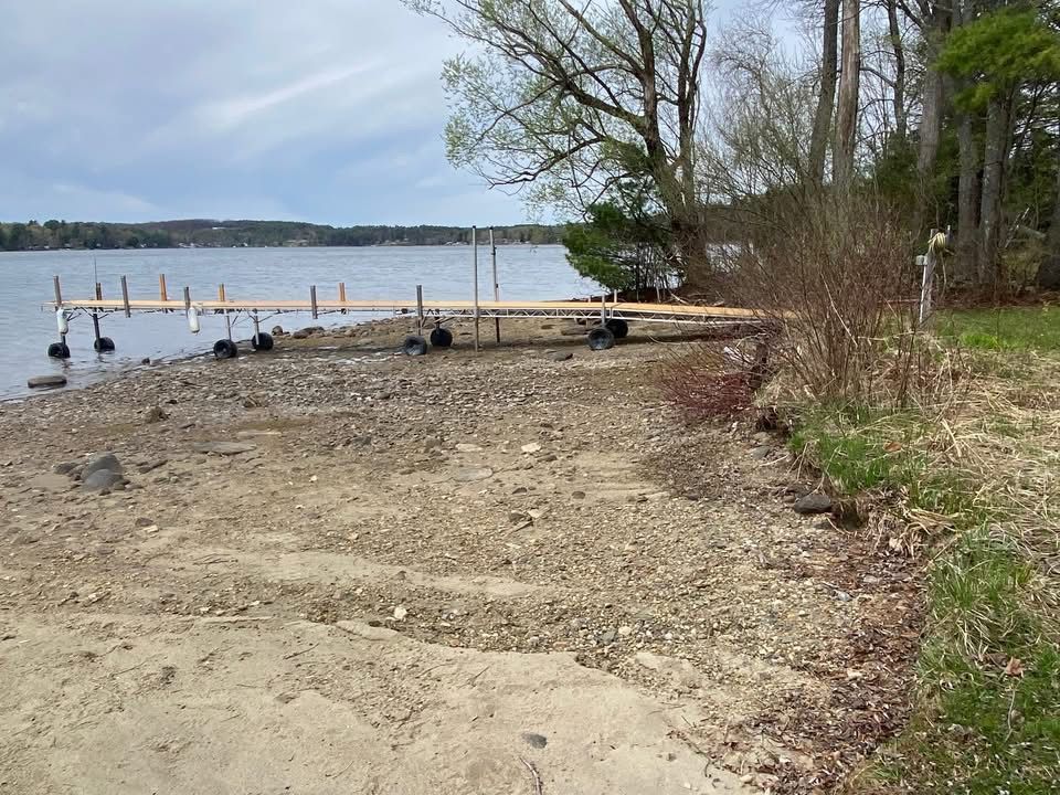 Rocky lakeshore with a wooden dock, leafless trees, and calm water under a cloudy sky