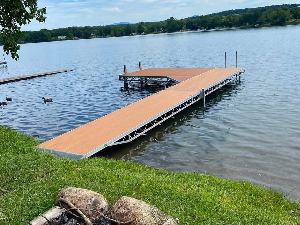 Floating wooden dock on a calm lake with grassy shore and distant trees under a clear sky