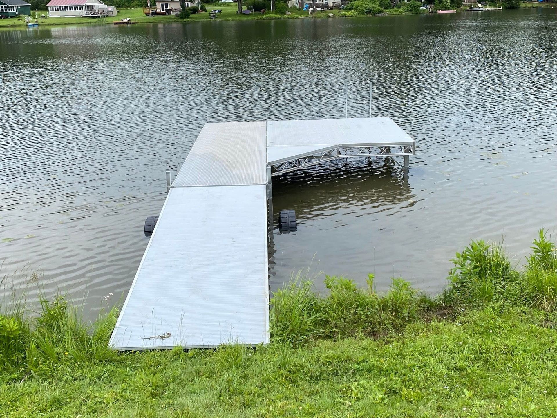 An L-shaped aluminum boat dock sitting in a calm lake near a grassy shoreline.