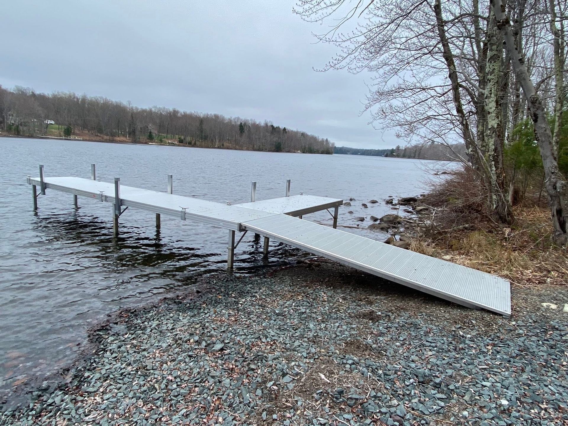 A white dock extends into a calm gray lake beside a rocky shore and leafless trees.
