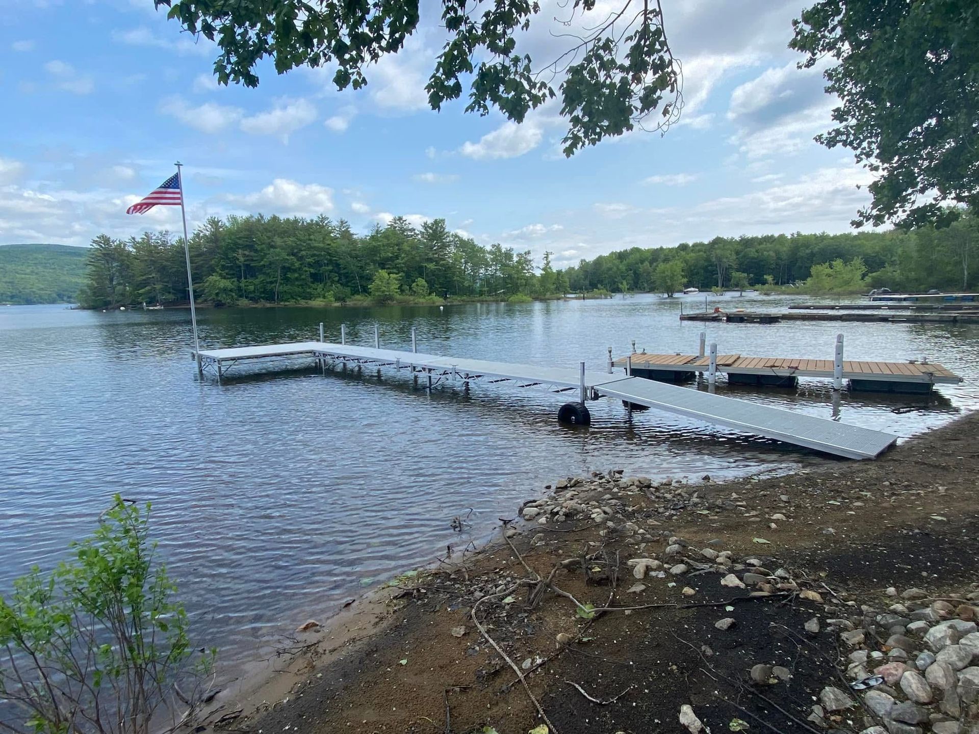 A metal dock with an American flag extends into a calm lake, with a wooded shoreline in the distance under a blue sky.