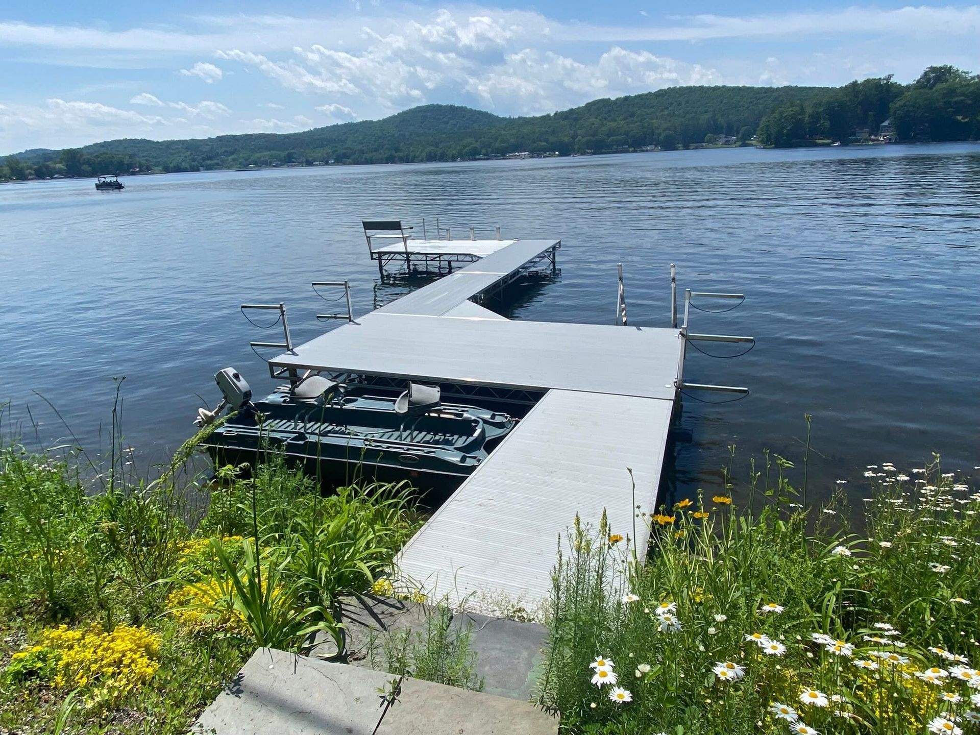A grey L-shaped dock extends into a calm lake, surrounded by green shore vegetation with mountains in the background.