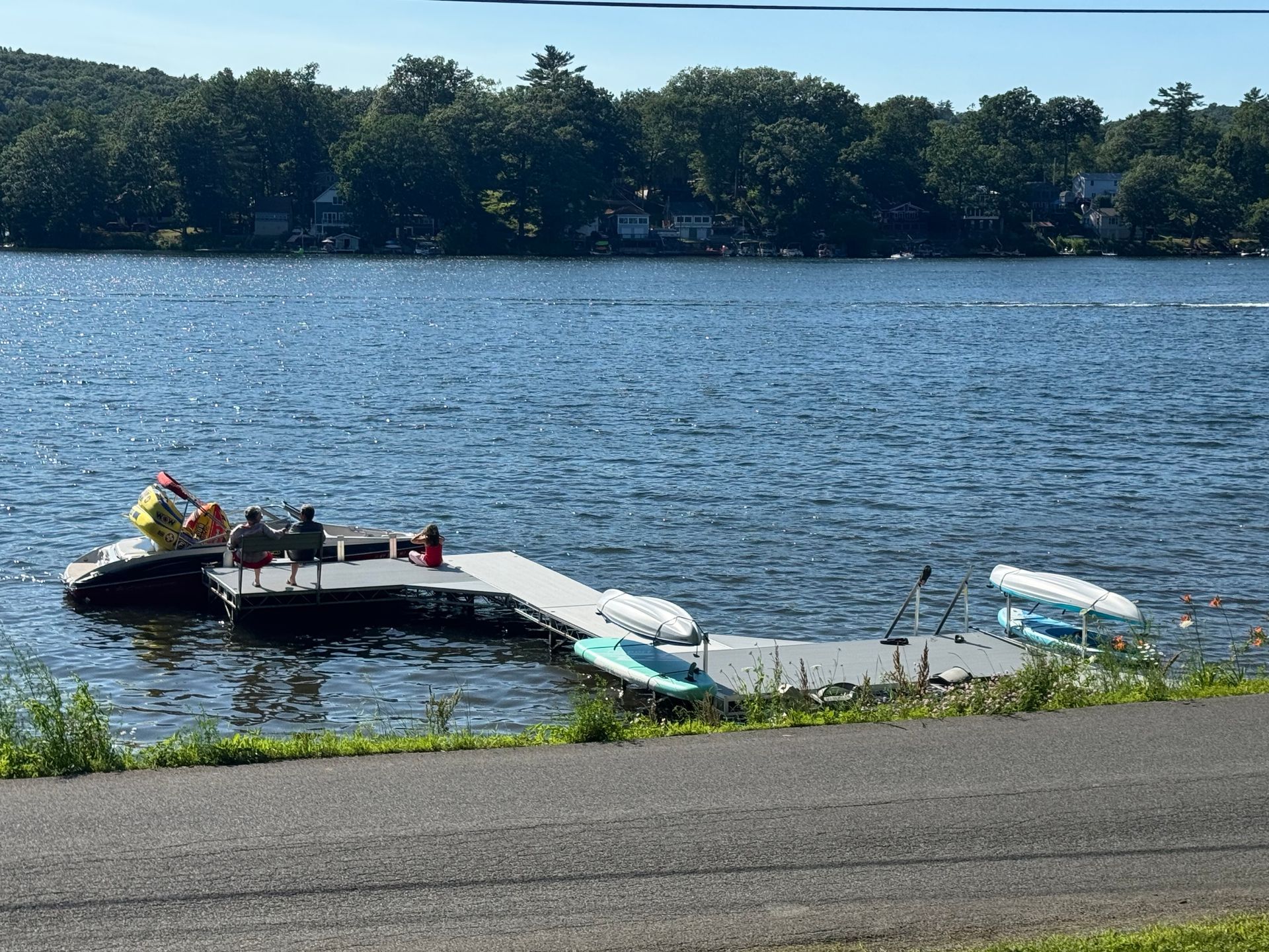 A floating dock on a lake with several people seated, kayaks and a paddleboard docked, and trees along the distant shore.
