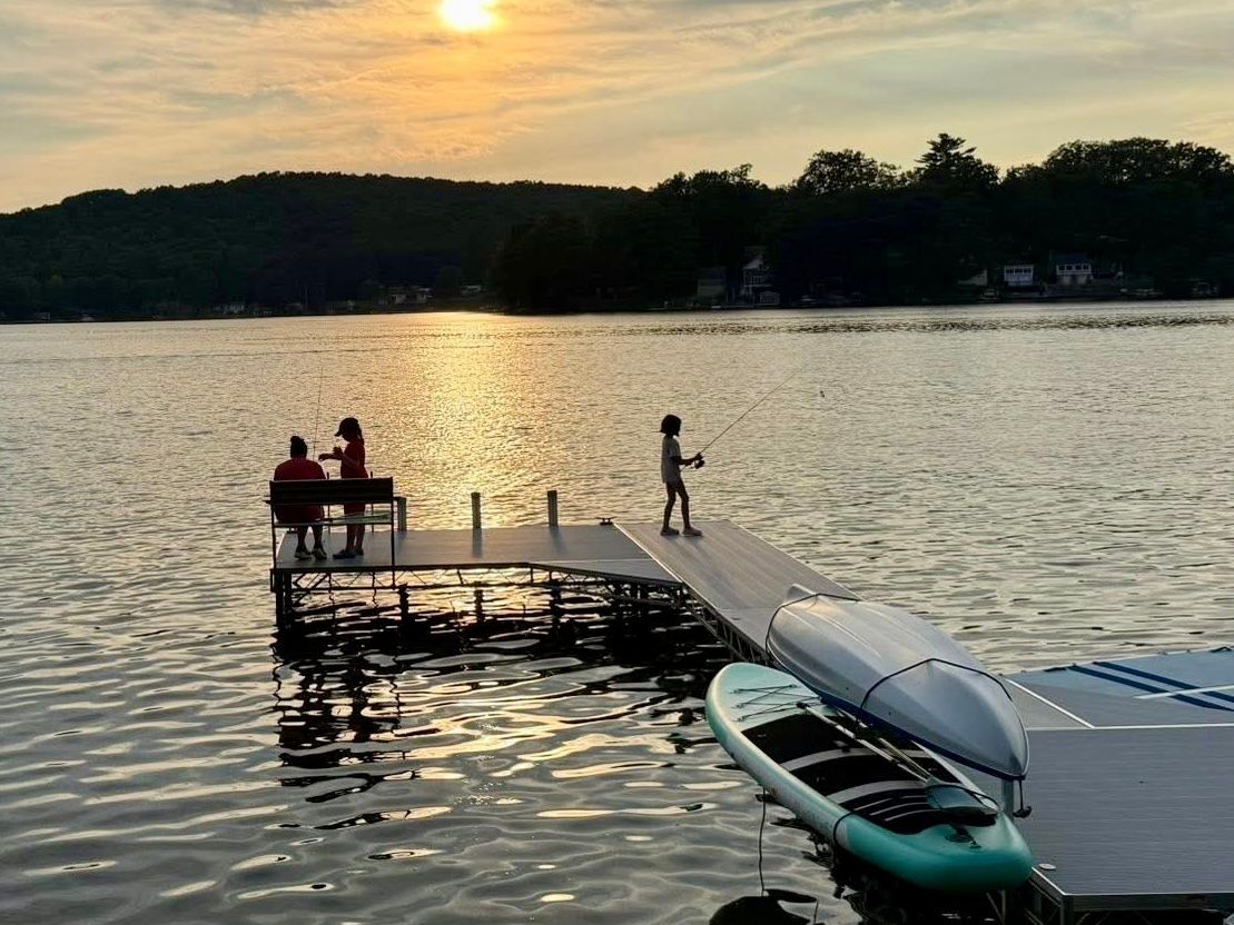 Three people fish from a wooden dock at sunset on a calm lake, with two kayaks resting on the edge nearby.