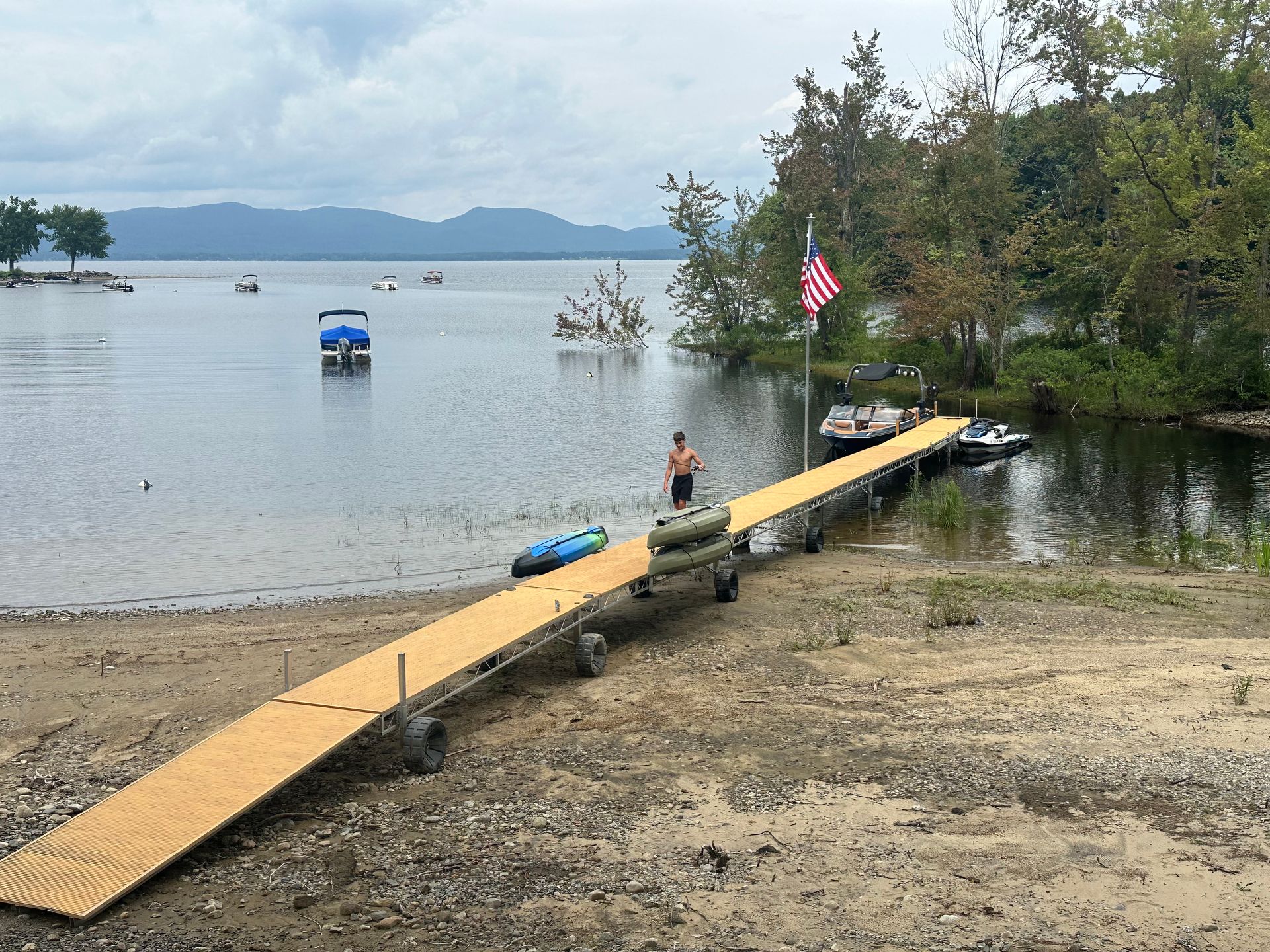 A wooden dock extends into a lake from a sandy shore, with a person, water tubes, and boats nearby under a cloudy sky.