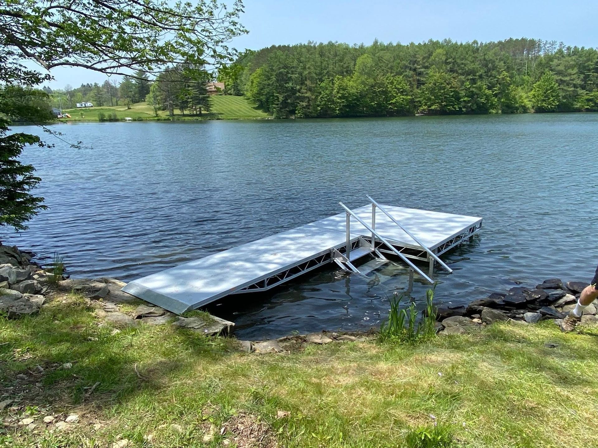 A silver dock with steps extends into a lake with trees and grass along the shore.