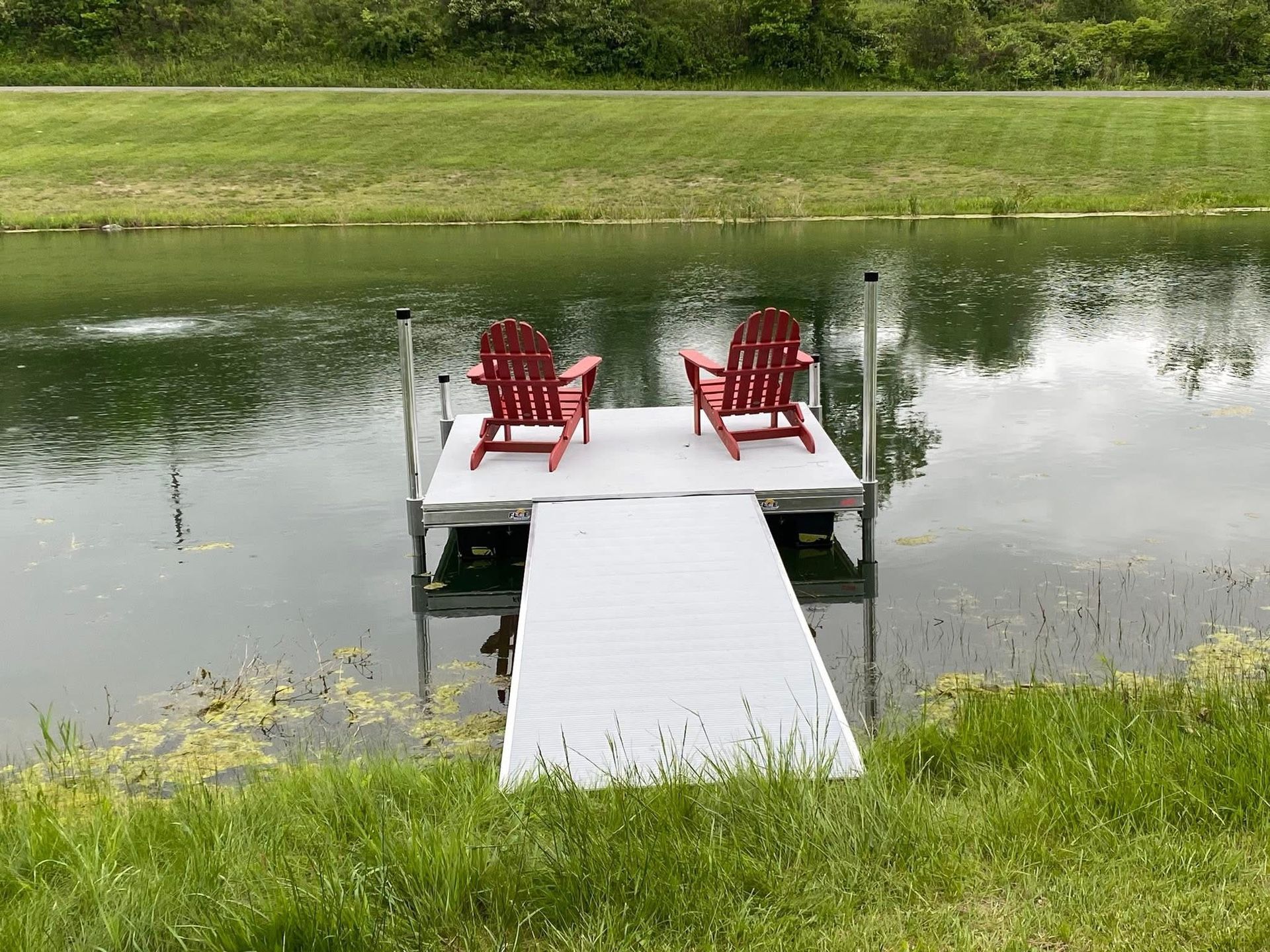Two red Adirondack chairs on a dock extending into a calm pond. Green grass surrounds the water.