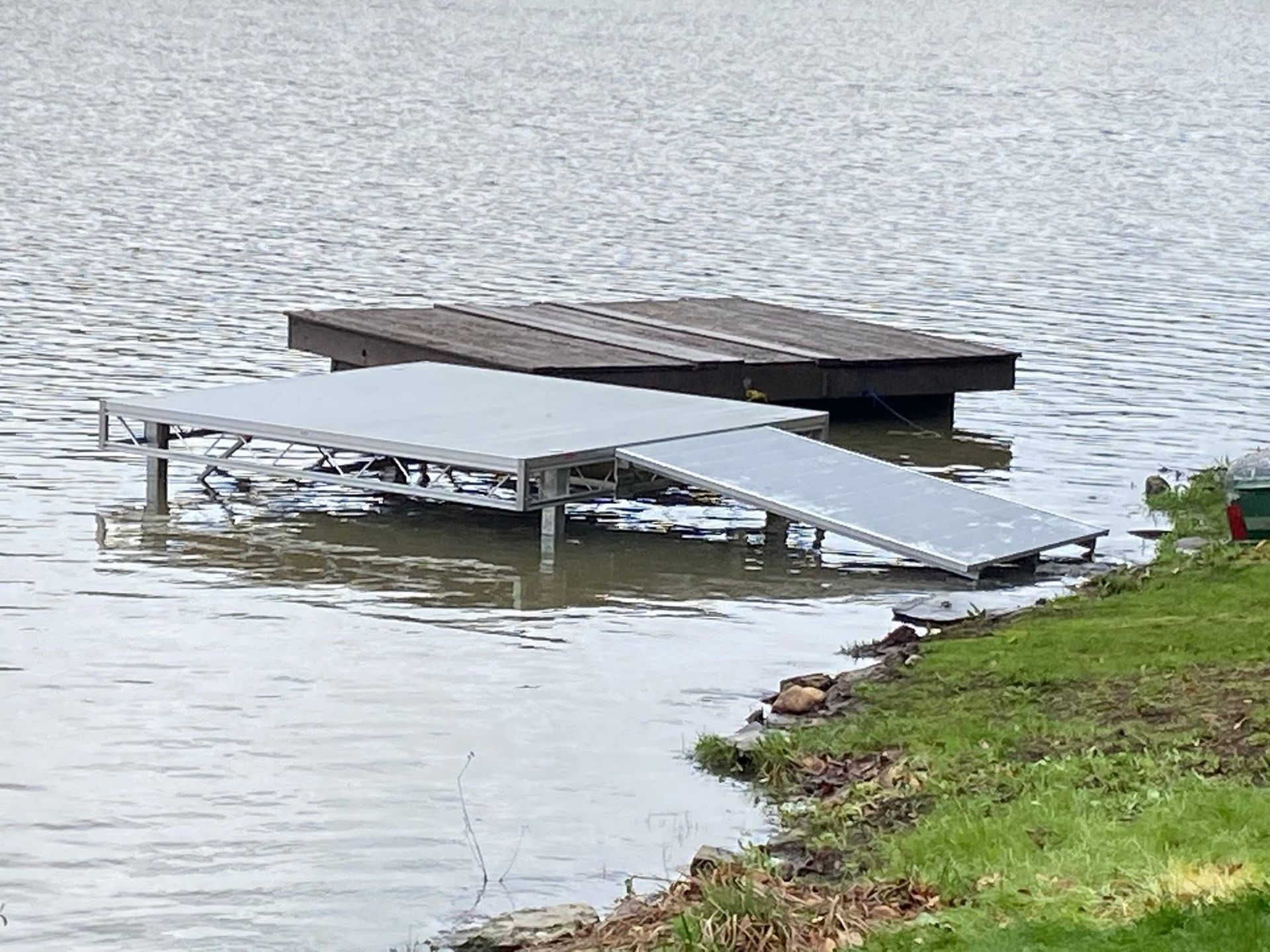 Dock with aluminum ramp and wooden platform in lake. Water level high, near grassy shore.