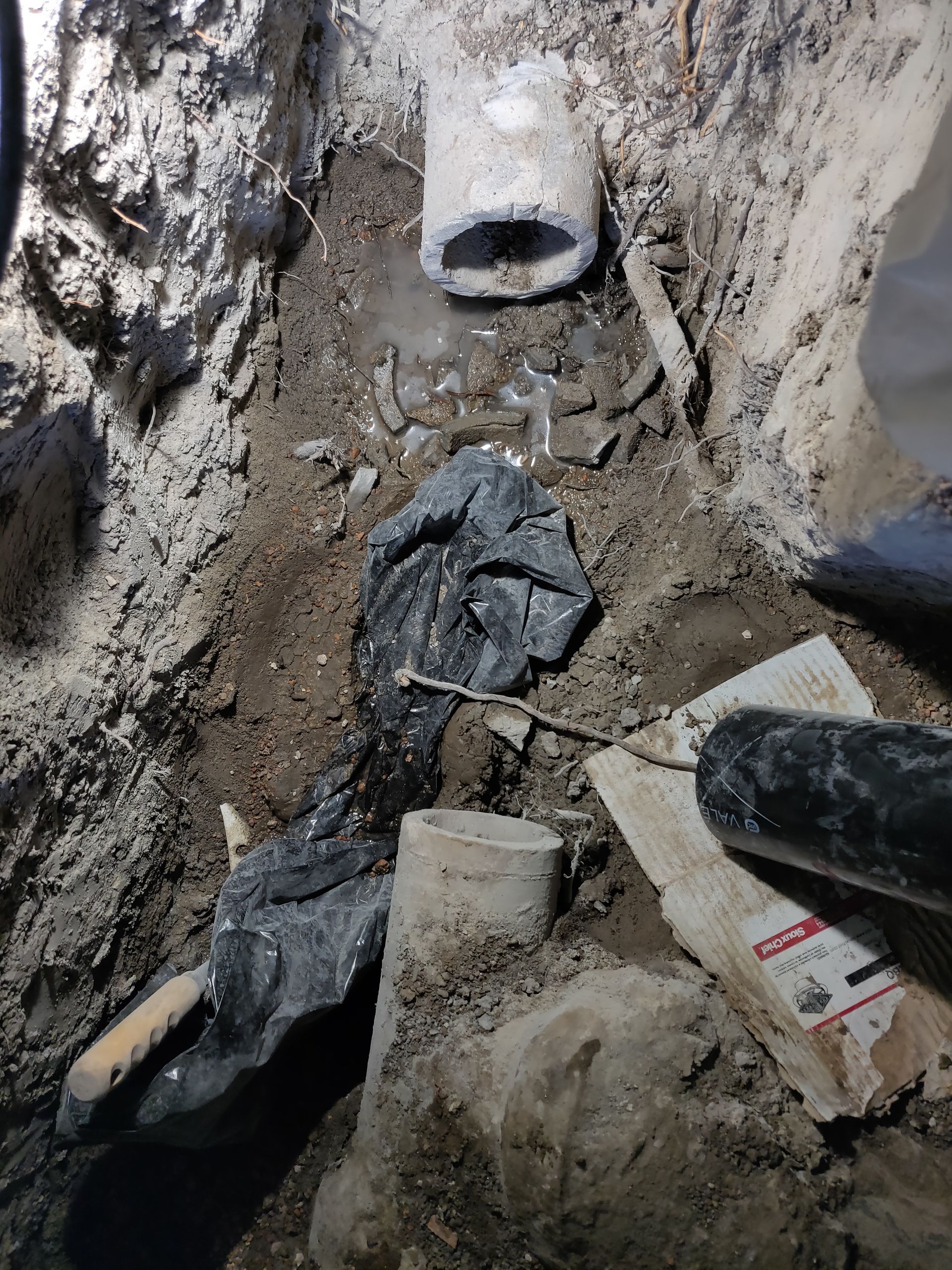 Excavated area showing plumbing pipes, black debris, and a trowel.