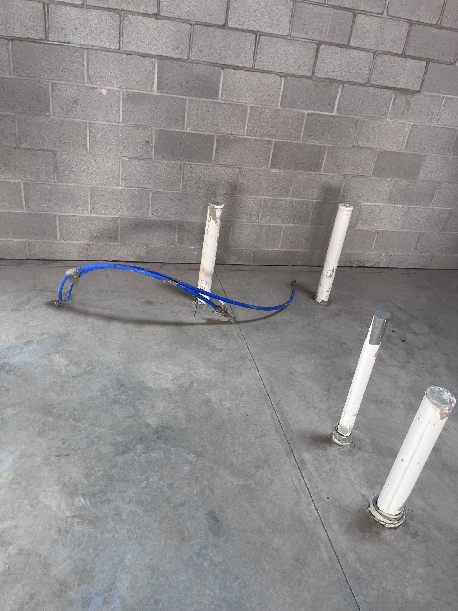 Concrete floor with exposed plumbing pipes and blue tubing against a cinder block wall.