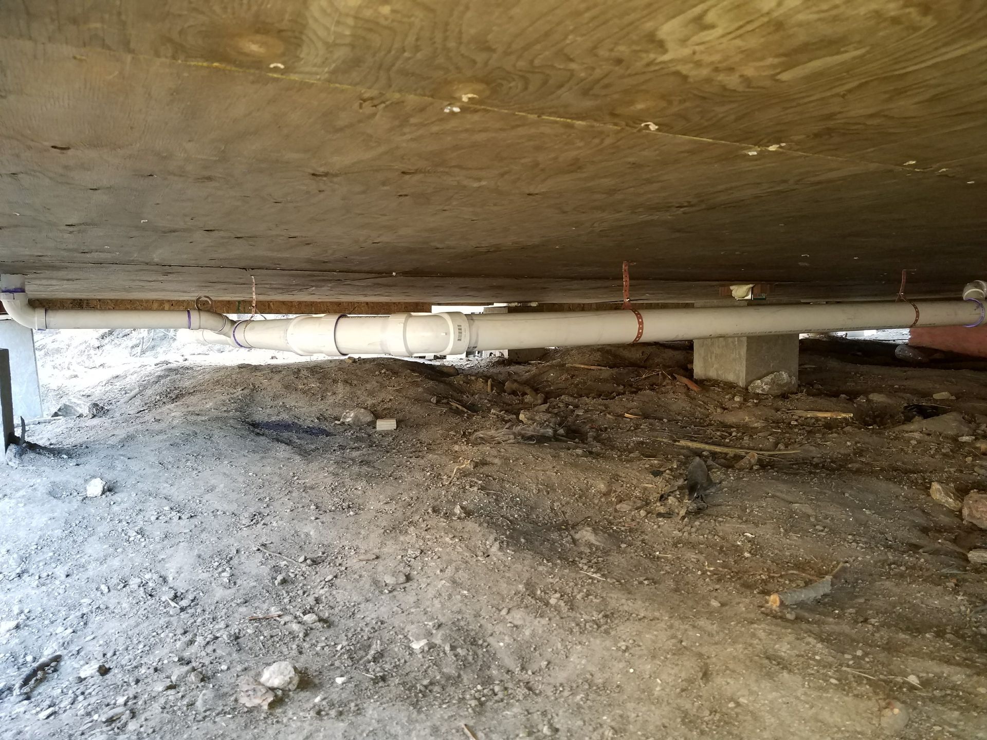 Underneath a structure, white pipes are suspended by wires, amid dirt and debris.