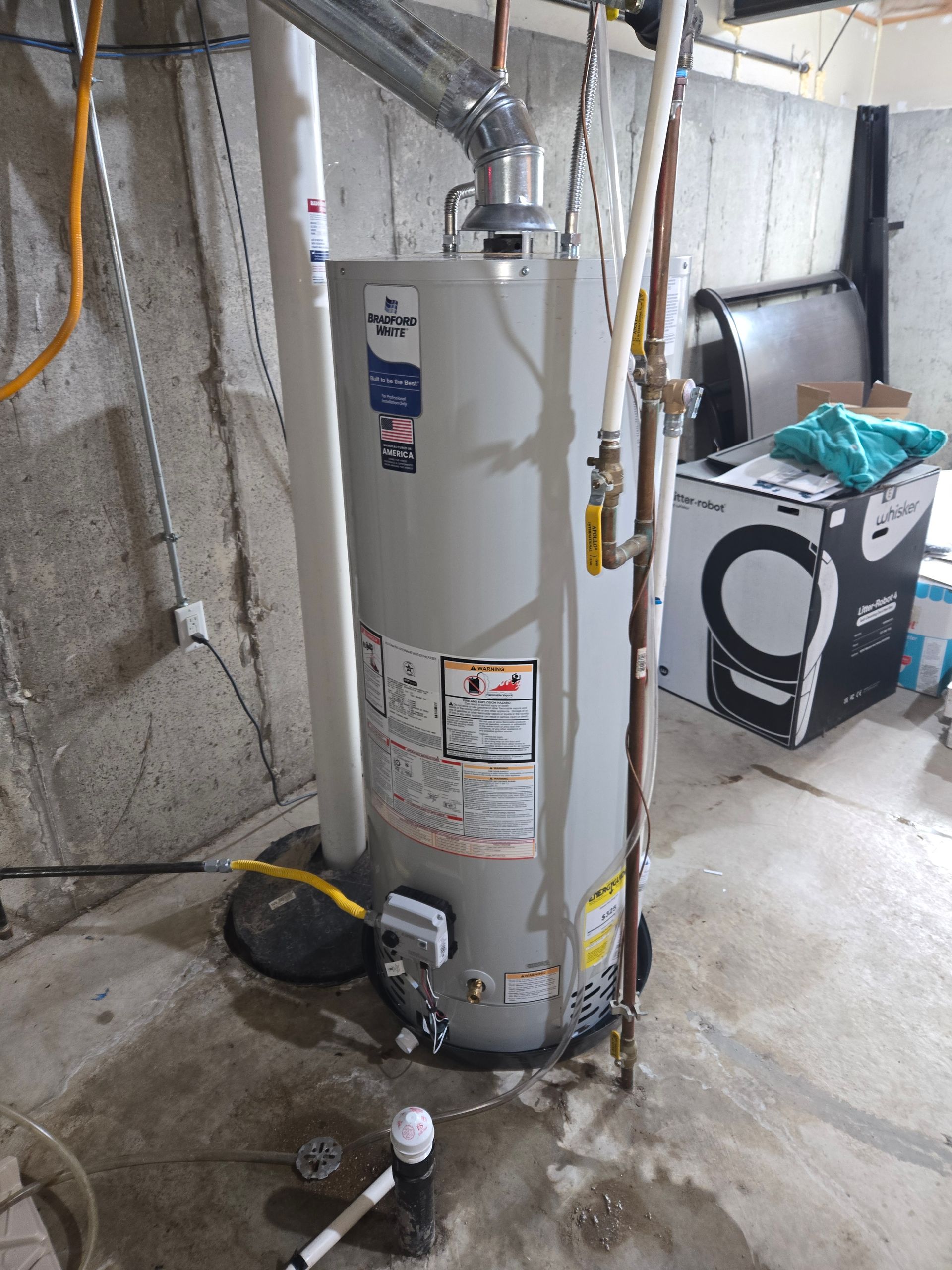 A gray water heater stands in a basement near pipes and a washing machine box.