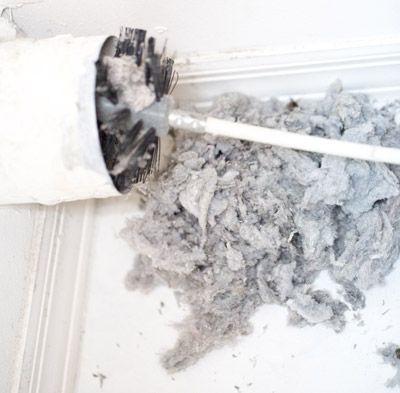 A dryer vent cleaning brush pulls a large pile of gray lint out of a white vent pipe.