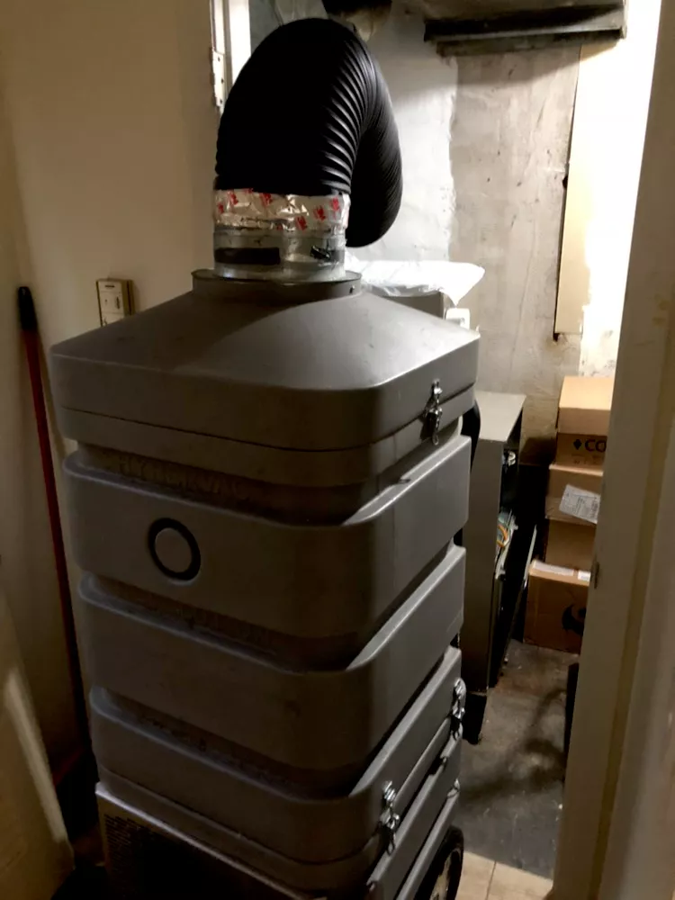 A grey, stacked air filtration unit with a black flexible vent hose stands in a dimly lit, cluttered basement corner.