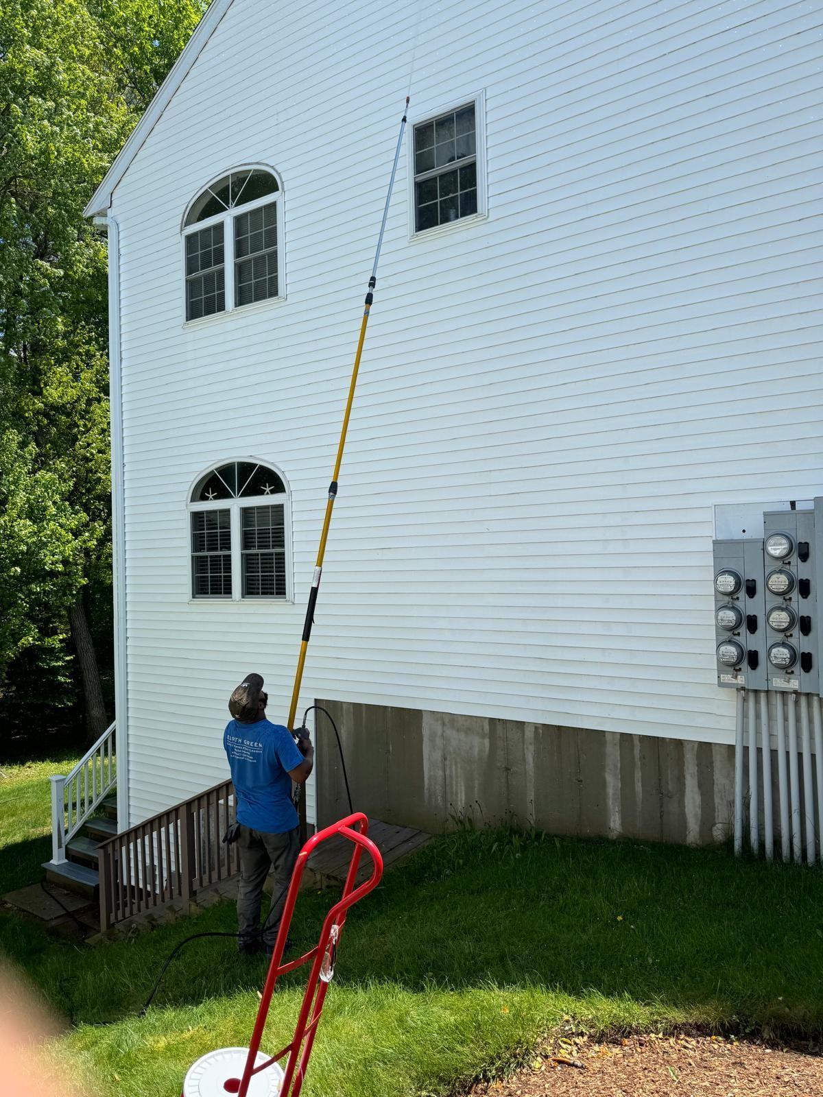 A worker uses an extended pole to clean the exterior siding of a white multi-story house near a utility box.