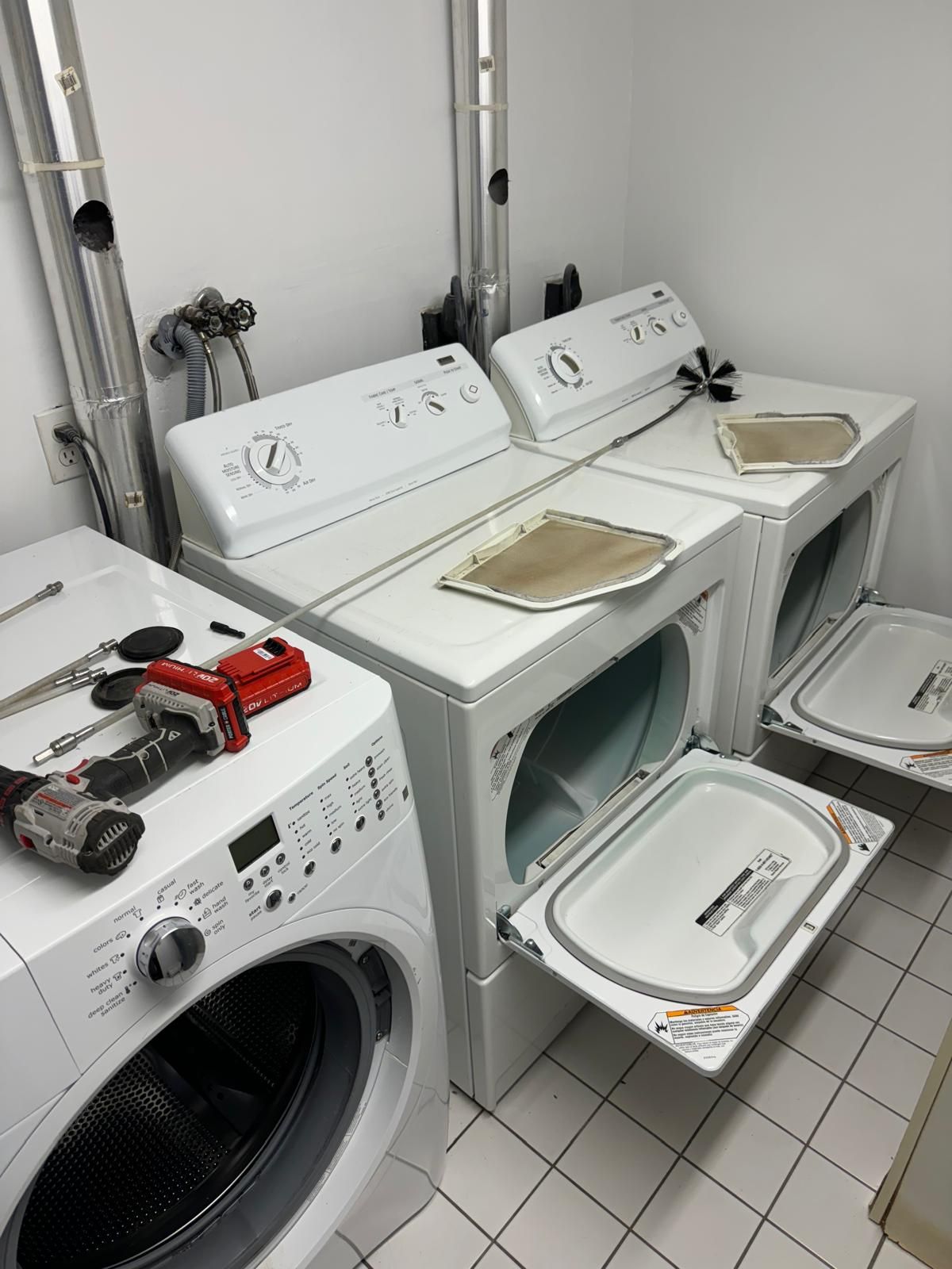 A modern front-load washer stands next to two open top-load dryers in a room with tiled floors and exposed plumbing.