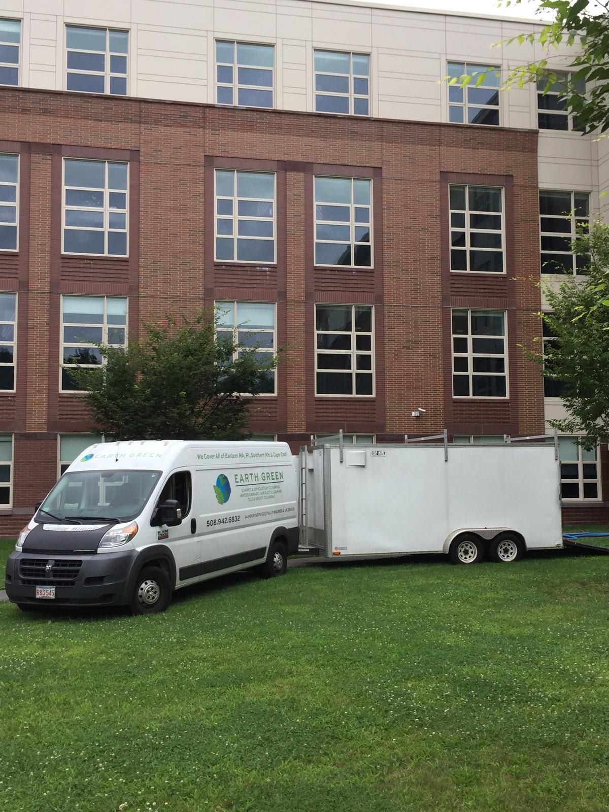 A white service van towing a matching white trailer parked on a grassy lawn in front of a brick building.