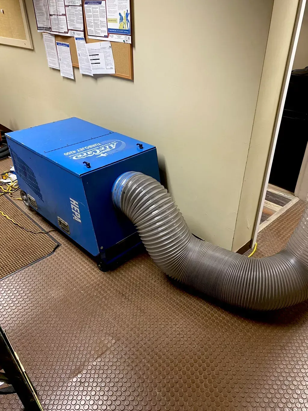 A blue industrial dehumidifier sits on a floor with a large, flexible grey hose extending from it into an adjacent room.